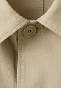 Beige overhemdkraag die bovenaan is dichtgeknoopt met fijne stiksels en een gladde plastic knoop die de kraag bevestigt.