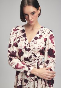 Dlouhé rukávy zavinovací šaty v krémové barvě s vínovým květinovým vzorem. Mají hluboký výstřih do V, nařasená ramena a zavázaný pas. Hladká textury látky.