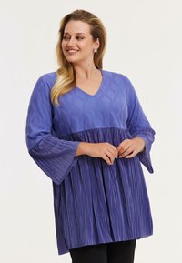 Lilla top med en tekstureret bodice, V-udskæring og plisserede klokkeslæt, parret med en plisseret nederste sektion.