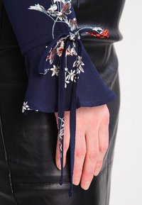 Ruka v černém koženém oděvu, tmavomodrý květinový rukáv s látkovou mašlí kolem zápěstí, ukazující detailní texturu a vzor.