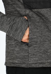 Giacca grigia con texture, dotata di una tasca sul lato sinistro con un pannello nero nella parte superiore. Il tessuto sembra elastico e morbido.
