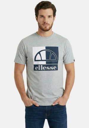 Homme barbu portant un t-shirt gris Ellesse et un jean bleu, debout les mains dans les poches devant un fond uni.