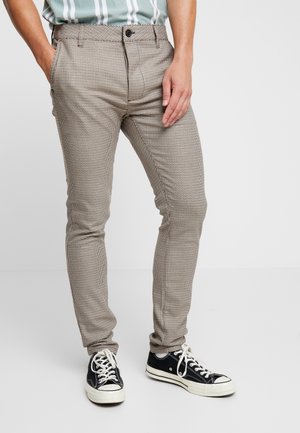 Mann trägt braune und weiße karierte Slim-Fit-Hose mit schwarzen und weißen Sneakers, steht mit einer Hand in der Hosentasche.