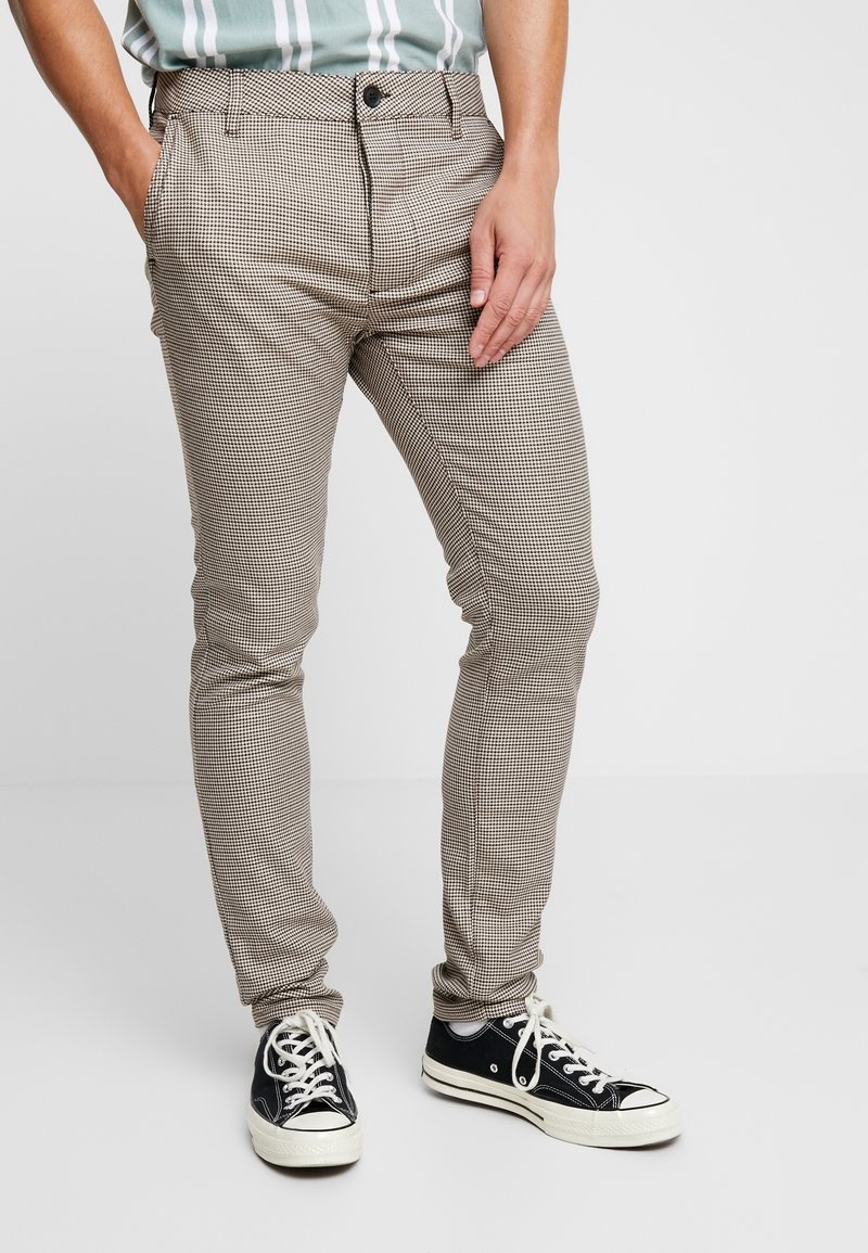 Homme portant un pantalon slim écossais marron et blanc avec des baskets noires et blanches, debout avec une main dans la poche.