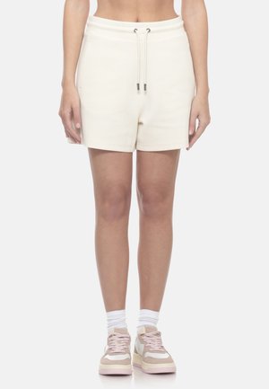 Donna che indossa pantaloncini color crema con coulisse, calzini bianchi e scarpe da ginnastica beige e bianche, in piedi davanti a uno sfondo semplice.