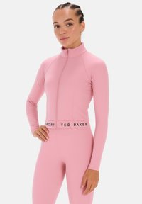 Világos rózsaszín sportpóló magas gallérral és teljes cipzárral, hosszú ujjal és logós szalaggal a deréknál. Simító, rugalmas anyag.