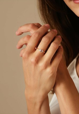 Roségoldring mit einem zentralen klaren Stein, gehalten zwischen zwei leicht strukturierten Händen vor einem sanften, neutralen Hintergrund.