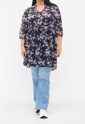 Vrouw met een marineblauwe tuniek met roze bloemenprint, lichtblauwe spijkerbroek en witte sneakers, staand tegen een effen witte achtergrond.