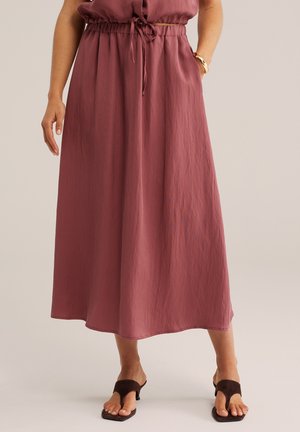 Vrouw die een mauve midi-rok draagt met een trekkoord in de taille, gecombineerd met donkerbruine open-teen sandalen met hak en een gouden armband om de pols.