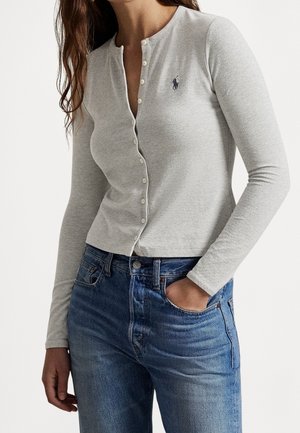Femme portant un cardigan gris clair à manches longues boutonné avec un petit logo brodé et un jean bleu, une main dans la poche.