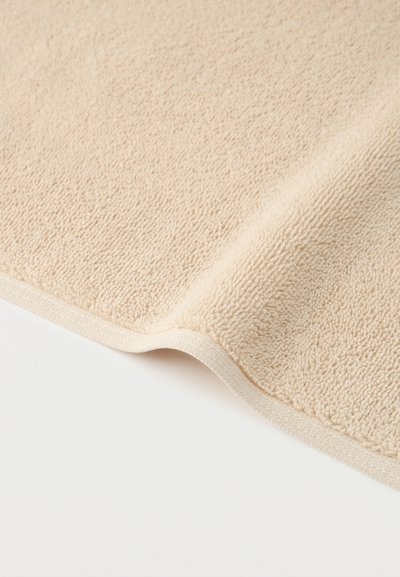 Μαλακό ύφασμα πετσέτας σε κρεμ απόχρωση με υφή από θηλιές και περιμετρική ραφή, τοποθετημένο σε λευκή επιφάνεια.