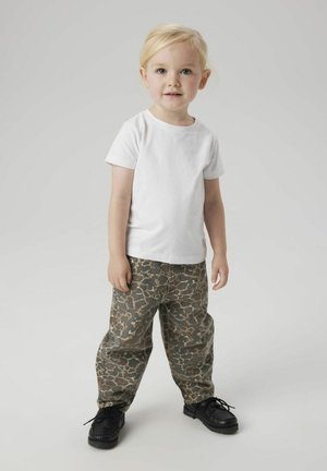 Laps, kes kannab valget lühikeste varrukatega T-särki, mustrilisi saapapükse rohelise ja pruuni tooni, ning musti seotud jalanõusid.
