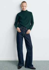 Maglione verde bosco a collo alto, abbinato a jeans scuri a gamba larga con allacciatura frontale e semplici scarpe nere con tacco.