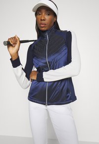 Námořnický proš�ívaný vest s bílými a námořnickými dlouhými rukávy, s obousměrným zipem a sportovním designem, držený s golfovým tičím na neutrálním pozadí.