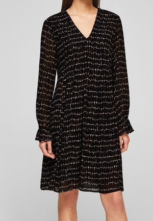 Žena v černých šatech ke kolenům s béžovým mřížkovaným vzorem, dlouhými rukávy, výstřihem do V a nařasenými manžetami.