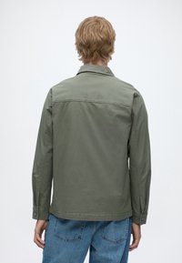 Personne aux cheveux châtain clair portant une chemise à manches longues vert olive et un jean bleu, vue de dos sur un fond clair uni.