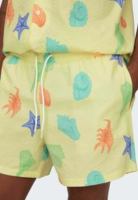 Shorts amarillos texturizados que presentan una variedad de patrones de conchas marinas y estrellas de mar en colores naranja, azul y verde. Cintura con cordón ajustable.