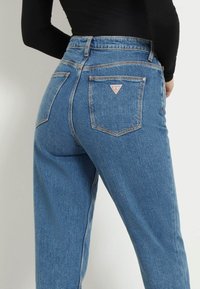 Persona que lleva unos vaqueros de cintura alta de mezclilla azul con un logo de Guess en el bolsillo trasero y una camiseta de manga larga negra, mostrado de la cintura hacia abajo.