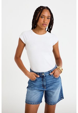 Ung kvinde med flettet hår iført en hvid tætsiddende T-shirt, blå denimshorts og gyldne armbånd, stående med hænderne i lommerne.