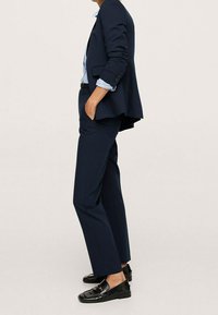 Osoba ubrana w granatowy garnitur z podwiniętymi rękawami, pasiastą koszulę i czarne mokasyny, stojąca z rękami w kieszeniach na jednolitym tle.