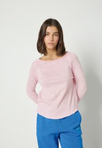 Mladá žena s hnědými vlasy po ramena, oblečená v světle růžovém tričku s dlouhým rukávem a modrých kalhotách, stojící před jednobarevným bílým pozadím.