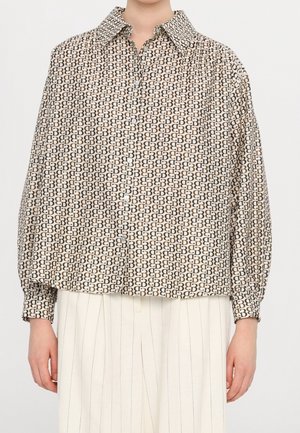 Personne portant une chemise à manches longues avec un motif géométrique noir et beige, associée à un pantalon blanc à fines rayures.