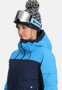 Blaue und marineblaue isolierte Skijacke mit hohem Kragen, Fronttaschen und einer glänzenden silbernen Skibrille auf einer navyblauen gemusterten Strickmütze.