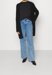 Svart lång cardigan, figursydd svart topp, högmidjade ljusblå vida jeans och svarta spetsiga stövlar. Släta, mjuka texturer.