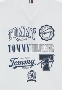 Hvit hettegenser med ulike marineblå logoer, inkludert "TOMMY," "Hilfiger," og et sirkulært emblem med "Sports Club New York siden 1985."