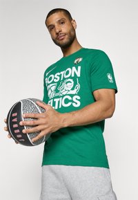 Zielona bawełniana koszulka z grafiką i logo "BOSTON CELTICS"; szare szorty; osoba trzymająca czarno-szary teksturowany kosz. 