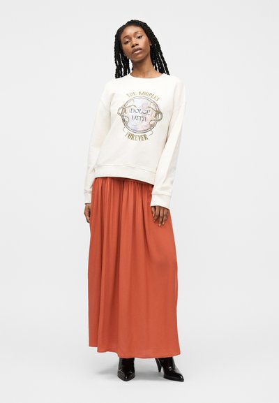 Γυναίκα με μακριά κοτσίδια που φοράει λευκή φούτερ με χρυσή εκτύπωση και ρέουσα μάξι φούστα σε χρώμα σκουριάς, στέκεται με μαύρα ψηλοτάκουνα μποτάκια.