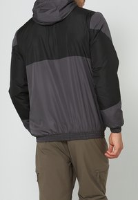 Giacca con cappuccio nera e grigia con una texture liscia e lucida. Presenta una vita elastica, pannelli sulle spalle a contrasto e una vestibilità comoda.