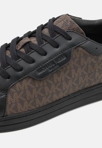 Baskets en cuir noir et tissu à motifs avec des lacets sombres et une semelle en caoutchouc. Présente la marque Michael Kors sur le côté.