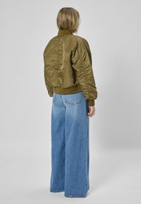 Olivgrüne Bomberjacke mit gerippten Bündchen und Saum, die eine geraffte Textur entlang des Rückens aufweist, kombiniert mit hoch taillierten, weit geschnittenen blauen Jeans.