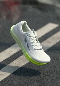 Λευκό και λαχανί αθλητικό παπούτσι με επάνω μέρος από δίχτυ και λογότυπο στο πλευρό, αιωρούμενο πάνω από άσφαλτο με λευκές οδικές γραμμές.