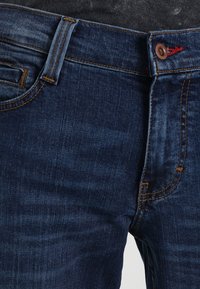 Jeans de denim azul oscuro con costuras naranjas contrastantes, que presentan un diseño simple, bolsillos frontales y un cierre de un solo botón.