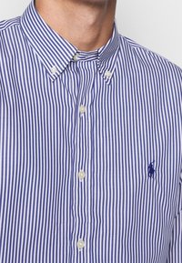 Mies, der ein blau-weiß gestreiftes Hemd mit Knopfkragen und gesticktem Polo-Spieler-Logo auf der Brust trägt.