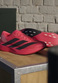 Φωτεινά ροζ παπούτσια για τρέξιμο Adidas με μαύρες ρίγες, κορδόνια και λεπτομερείς σόλες, τοποθετημένα πάνω σε ξύλινη επιφάνεια μπροστά από ένα λευκό τοίχο από τούβλα.