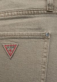 Bézs színű farmeranyag varrási részletekkel. A hátsó zsebén piros és fehér háromszög logócímke található. Fém szegecsek díszítik.