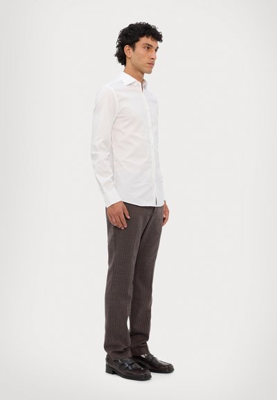 Witte overhemdd met kraag, gecombineerd met bruine pinstripe broeken en zwarte instappers. De stof lijkt soepel en aansluitend.