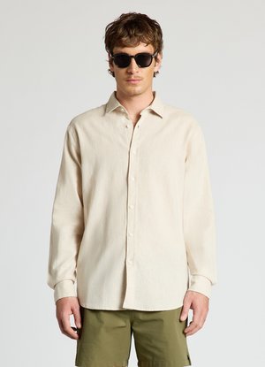 Mann trägt schwarze Sonnenbrille, beige langärmeliges Hemd mit Knöpfen und olivgrüne Shorts, steht vor einem schlichten hellgrauen Hintergrund.
