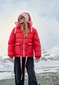 Czerwona puchowa kurtka z różowymi akcentami i dwoma przednimi kieszeniami, z regulacją w talii na sznurek, noszona na czarnych spodniach na tle zaśnieżonego krajobrazu.