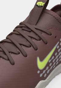 Krupni plan smeđe atletske cipele sa smeđim vezicama, neonsko žutim Nike logom, reflektirajućim točkastim uzorkom i oznakom Air na petlji jezika.