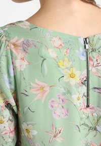 Grön blommig blus med ett färgglatt mönster av olika blommor, synlig dragkedja bak och mjukt, texturerat tyg.