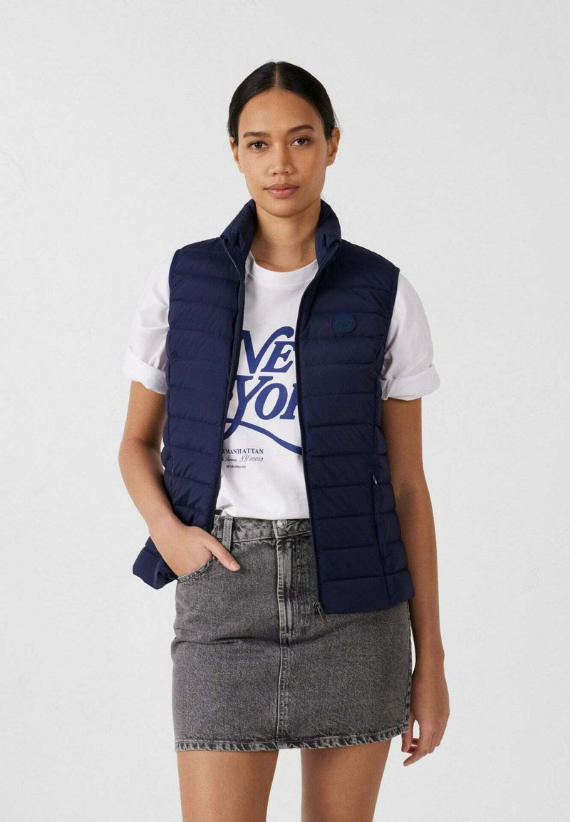 Veste matelassée bleu marine avec col montant, design matelassé et détail logo, portée sur un T-shirt blanc et une jupe en denim gris.