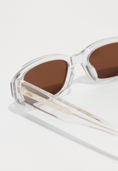 Clear plastic sunglasses with brown lenses, angular frame design, and textured gold-accented temples. Light and sleek appearance.