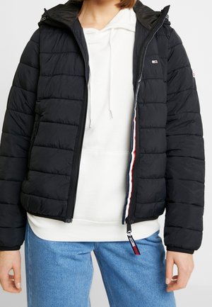 Osoba nosząca czarną pikowaną kurtkę na białej bluzie z kapturem oraz niebieskie dżinsy, prezentująca przedni zamek błyskawiczny i detale z logo na kurtce.