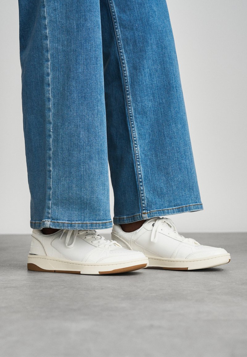 White leather sneakers with a smooth texture, lace-up design, and brown rubber sole, paired with cropped, wide-leg blue denim jeans.