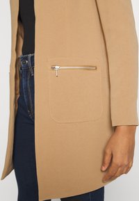 Beige texturerad kappa med en sidofickapåse och silverfärgad dragkedja, kombinerad med mörkblå jeans.