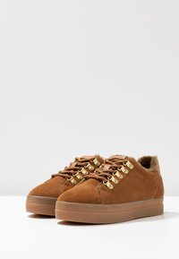 Baskets en suede marron avec des lacets en métal doré, doublure douce et une semelle en caoutchouc épaisse et texturée. Bout arrondi et design bas.